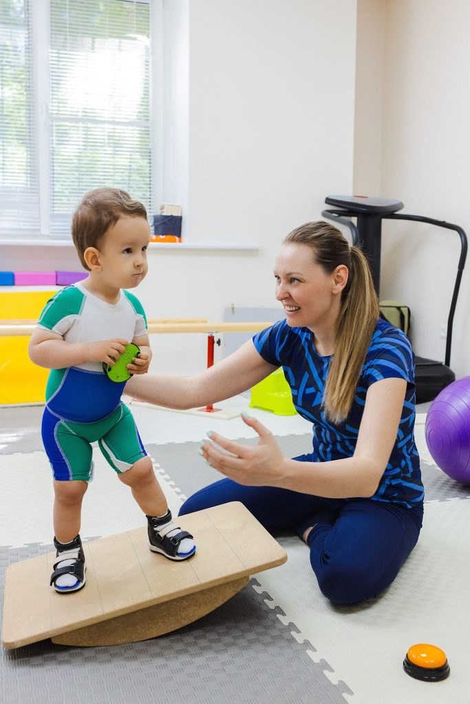 A smiling female pediatric physiotherapist sitting on a foam mat and guiding a toddler boy wearing a specialized compression suit and leg orthotics as he balances on a wooden wobble board.