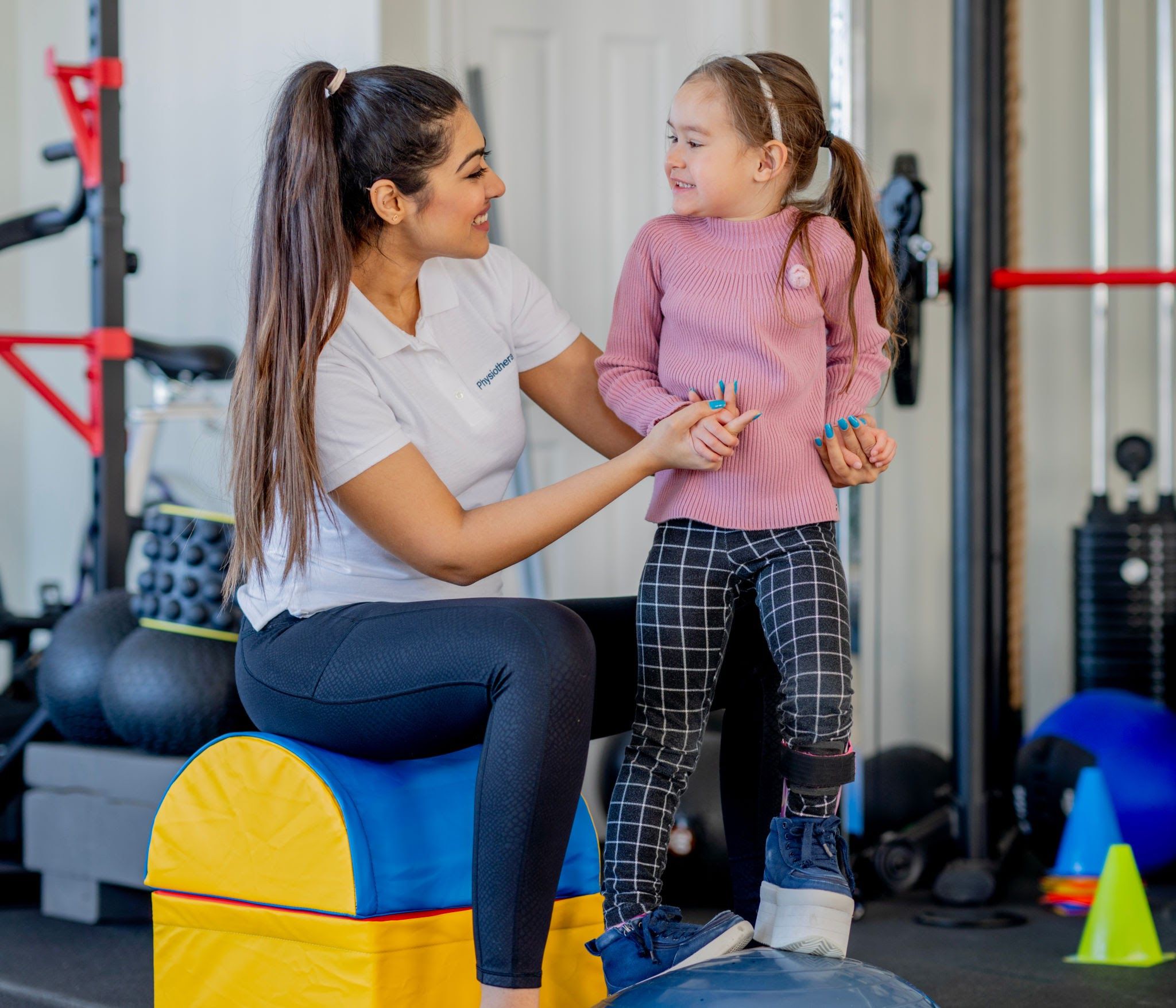 A smiling female pediatric physiotherapist assisting a young girl wearing a lower leg brace as she practices balance exercises on a blue stabilization ball.